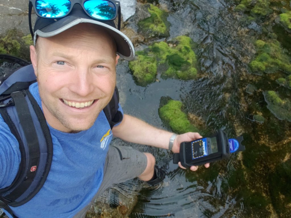 Person with hat and glasses holding geiger counter while wading in stream with hiking boots and backpack