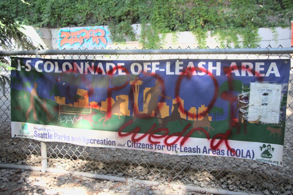 Big banner that says "I-5 Colonnade off-leash area" with "POOP WEDREA" spray painted over it in red